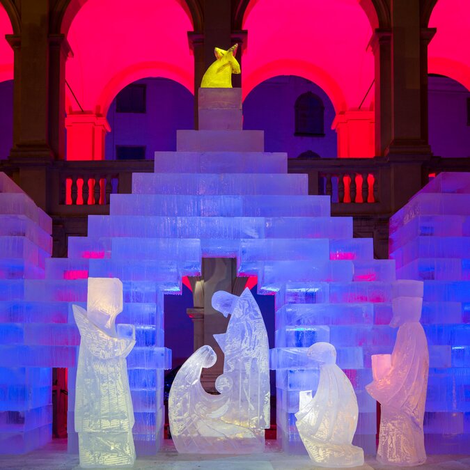 Illuminated ice sculpture depicting a nativity scene in front of a monumental installation made of ice blocks within a historic arcaded courtyard. Blue, purple, and pink lighting effects highlight the winter-themed artistic display. | © Graz Tourismus, Harry Schiffer