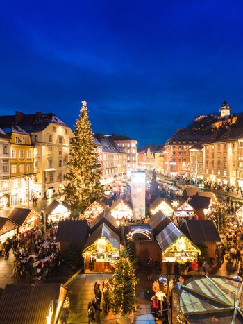 Marché de Noël illuminé sur une place historique à la tombée de la nuit. De nombreux chalets, un grand sapin décoré et de nombreux visiteurs créent une ambiance chaleureuse de l’Avent, tandis que les bâtiments environnants et la tour de l’horloge sont éclairés en arrière-plan. | © Graz Tourismus, Harry Schiffer