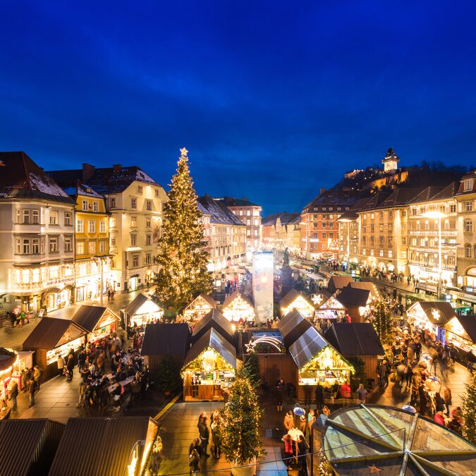 Festively illuminated Christmas market on a historic city square at dusk. Numerous market stalls, a large decorated Christmas tree and many visitors create a warm holiday atmosphere, while the surrounding buildings and the clock tower glow in the background. | © Graz Tourismus, Harry Schiffer