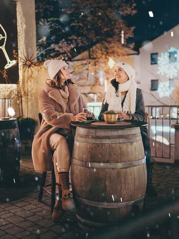 Deux personnes sont assises autour d’une table en forme de tonneau sur une terrasse chaleureusement éclairée en hiver. Des flocons de neige tombent tandis que des décorations lumineuses festives illuminent la vieille ville en arrière-plan. | © TVB Kufsteinerland, Thorben Jureczko