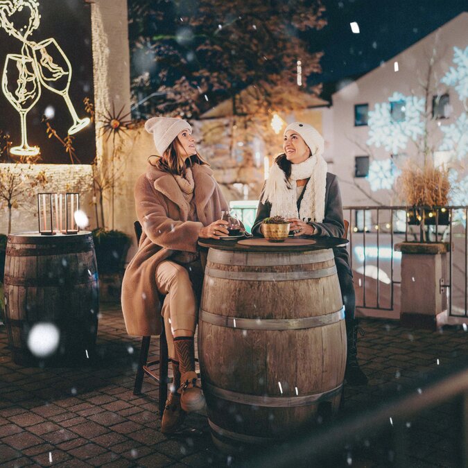 Two people sit at a barrel table on a warmly lit terrace during winter. Snowflakes fall as festive light decorations and seasonal displays brighten the historic town setting at night. | © TVB Kufsteinerland, Thorben Jureczko