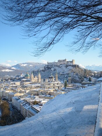 Vue panoramique hivernale sur Salzbourg avec la forteresse de Hohensalzburg, les toits enneigés et la rivière Salzach. Un sentier recouvert de neige longe la colline au-dessus de la vieille ville, bordé d’arbres dénudés et des Alpes en arrière-plan. | © Tourismus Salzburg GmbH