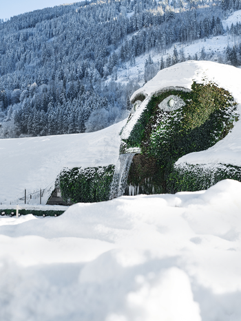 Paysage hivernal avec le célèbre Géant Swarovski à Wattens, dont le visage recouvert de végétation est partiellement enneigé. De l’eau s’écoule de la bouche de la sculpture, tandis que des collines et des forêts enneigées entourent le site. | © Swarovski Kristallwelten