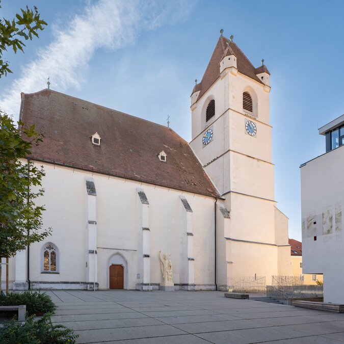 La cathédrale Saint-Martin d’Eisenstadt avec son clocher caractéristique et sa façade historique sobre. La place est entourée d’arbres et d’éléments d’architecture moderne sous un ciel bleu dégagé. | © Eisenstadt(c)Burgenland Tourismus_Andreas Hafenscher (45)
