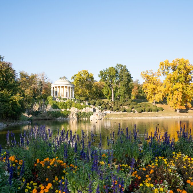 Le temple néoclassique de Léopoldine, situé dans le parc du château d’Eisenstadt, se dresse au bord d’un lac. Des massifs de fleurs colorées, des arbres aux teintes automnales et leurs reflets dans l’eau composent un paysage paisible. | © Leopoldinentempel_5(c)Burgenland Tourismus_Birgit Machtinger