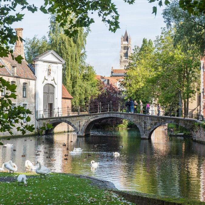 Stone bridge over the Minnewater in Bruges, surrounded by swans, trees, and historic houses. | © Piet de Kersgieter – Brügge, Blick auf Wijngaardbrug