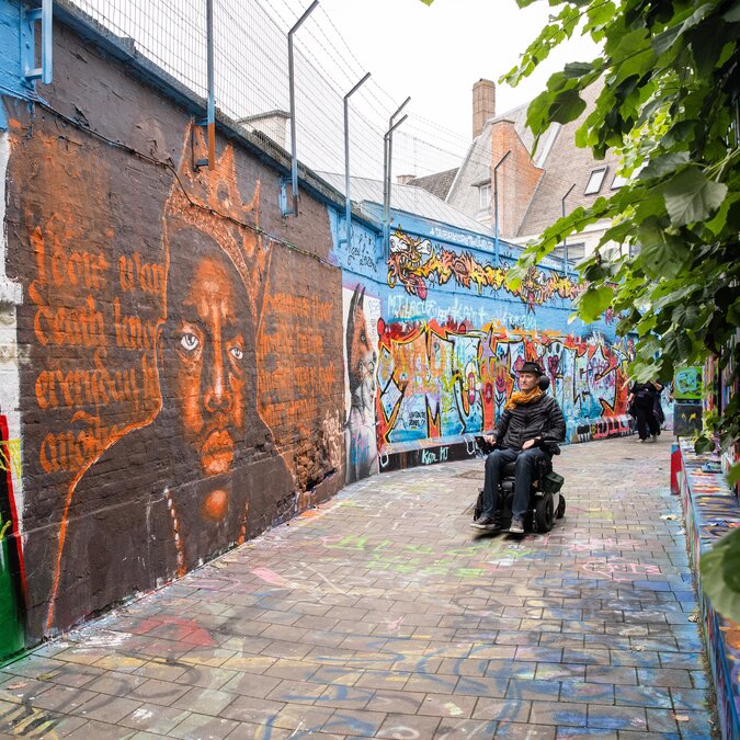 Graffitigasse in Gent mit farbenfrohen Wandmalereien und einer Person im elektrischen Rollstuhl. | © Piet De Kersgieter