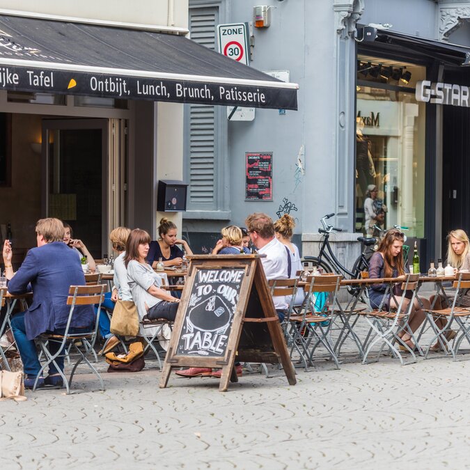 Belebte Terrasse eines Cafés in Gent mit Gästen an Holztischen auf einer Kopfsteinpflasterstrasse. | © Piet de Kersgieter – Terrasse in Gent