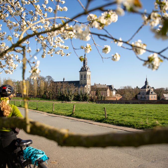 Person mit Fahrrad auf einer Landstrasse vor blühenden Obstbäumen, mit Blick auf eine historische Kirche und umliegende Felder in einer ländlichen Region. | © Stad Leuven