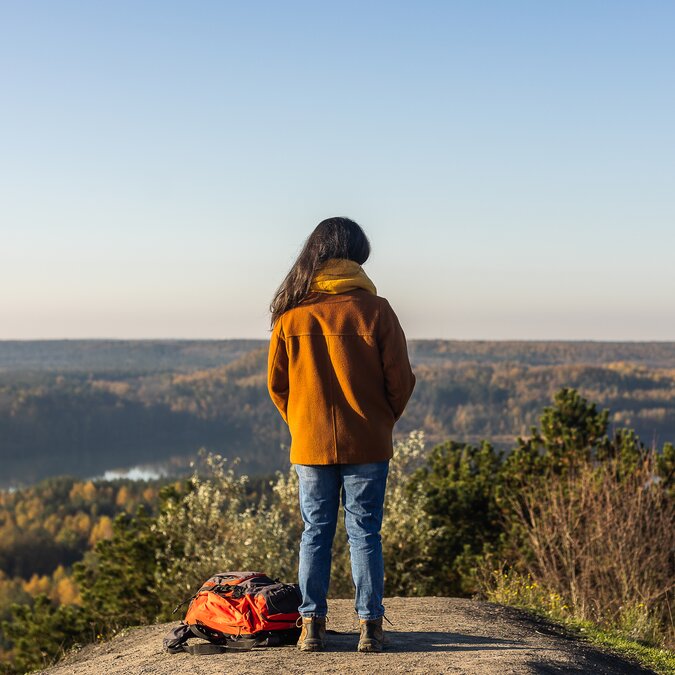 Une personne se tient sur un promontoire rocheux avec vue sur un vaste paysage composé de forêts et de lacs ; un sac à dos est posé par terre à côté d'elle. | © MEDIALIFE.BE