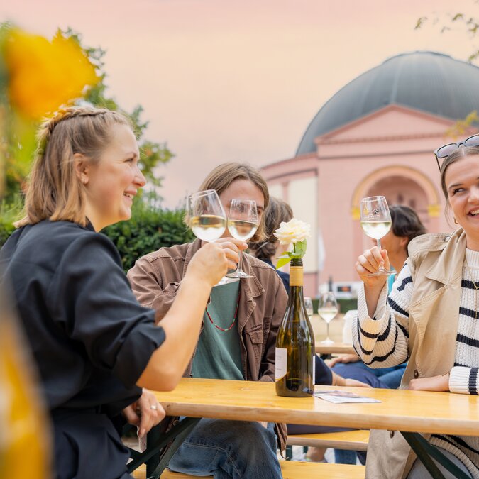 A group of people sit at a wooden table outdoors, raising glasses of white wine; in the background stands a pink church with a dome, surrounded by trees in the warm evening light. | © #visitrheinmain | David Vasicek