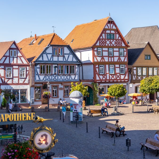 View of Seligenstadt’s market square with historic half-timbered houses, orange roofs and small trees. People sit on benches and at tables on the cobbled square in sunny weather. | © #visitrheinmain | David Vasicek