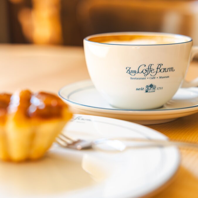 Coffee cup with the Coffe Baum Leipzig logo and pastries on a wooden table in warm light. | © Philipp Kirschner | Leipzig Travel