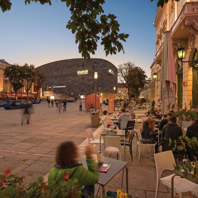 Blick auf einen Platz in der Abenddämmerung aus einem Café im Hintergrund das mumok und die Halle aus dem Museumsquartier in Wien.  | © Wien Tourismus | Christian Stemper