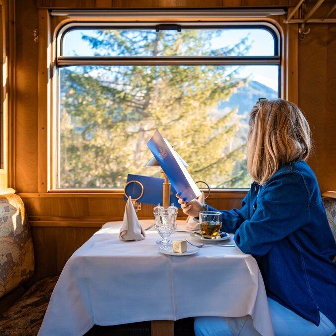 A person is sitting in the Gourmino, the dining car of the Glacier Pullmann Express, looking out of the window, holding a menu in her hand and a tea in front of her. | © Rhätische Bahn 