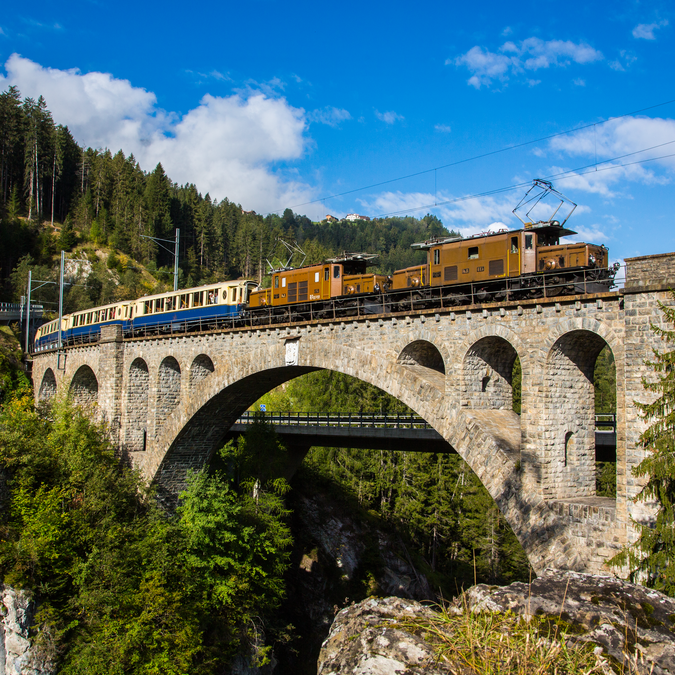 The Glacier Pullmann Express with a brown locomotive and white-blue carriages crosses a bridge in the middle of the forest, with the mountains and a blue sky behind it. | © Rhätische Bahn 