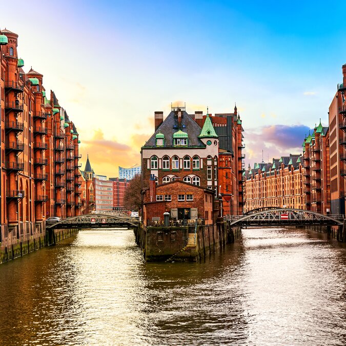 Le bâtiment historique du Wasserschloss, situé dans la Speicherstadt de Hambourg, se dresse entre deux canaux et est entouré d’anciens entrepôts en briques rouges. Des ponts franchissent les canaux et l’Elbphilharmonie est visible à l’arrière-plan. | © shutterstock_1946862949